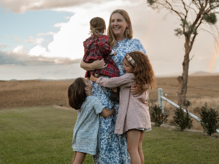 A mother holding a young child while her two daughters hug her around the waist, standing together in open countryside.