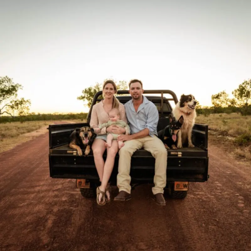 Family with their baby and working dogs on the back of a ute on a red dirt road