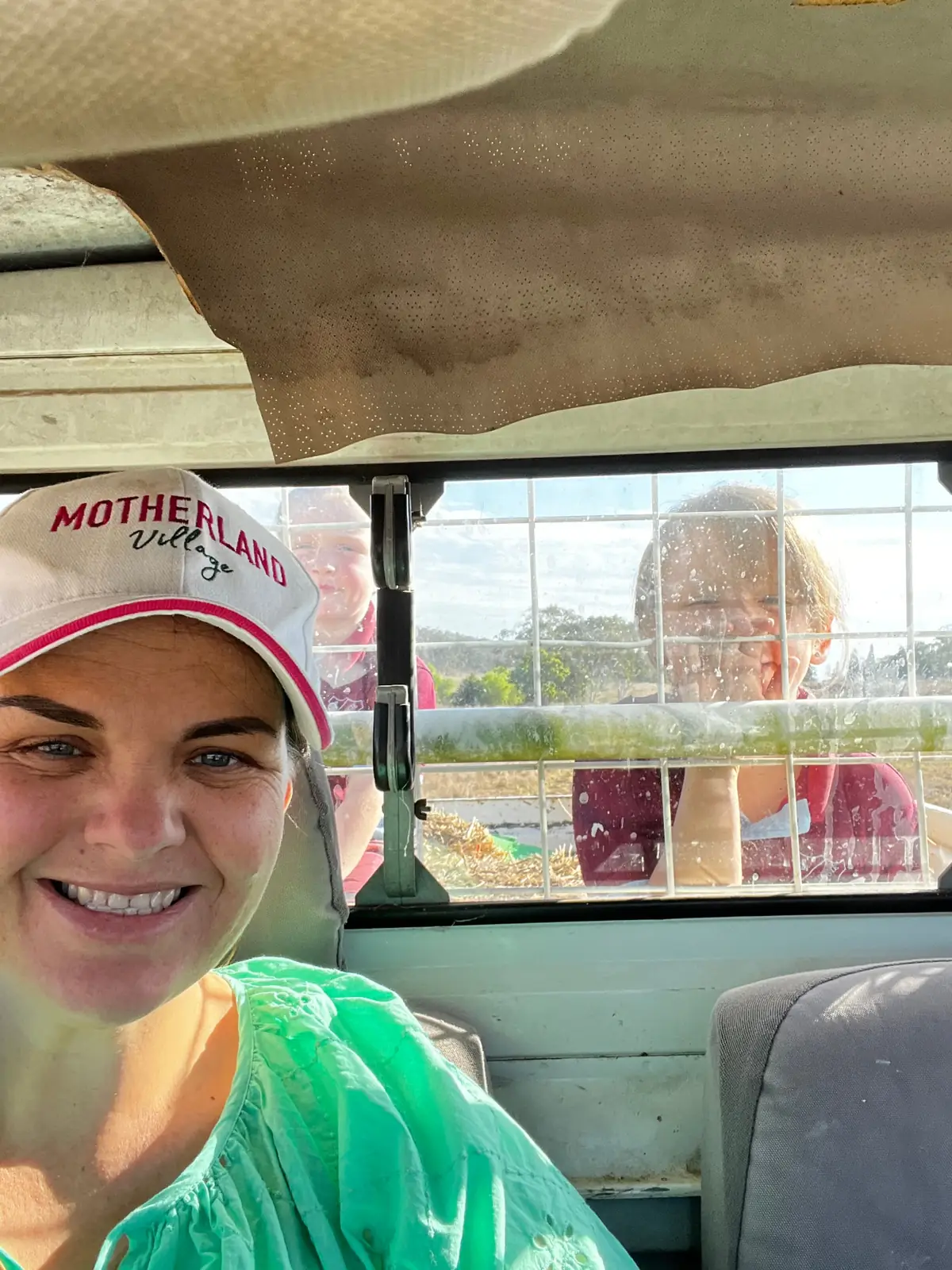 Mel smiling in her Motherland Village cap, with her kids behind her in the ute