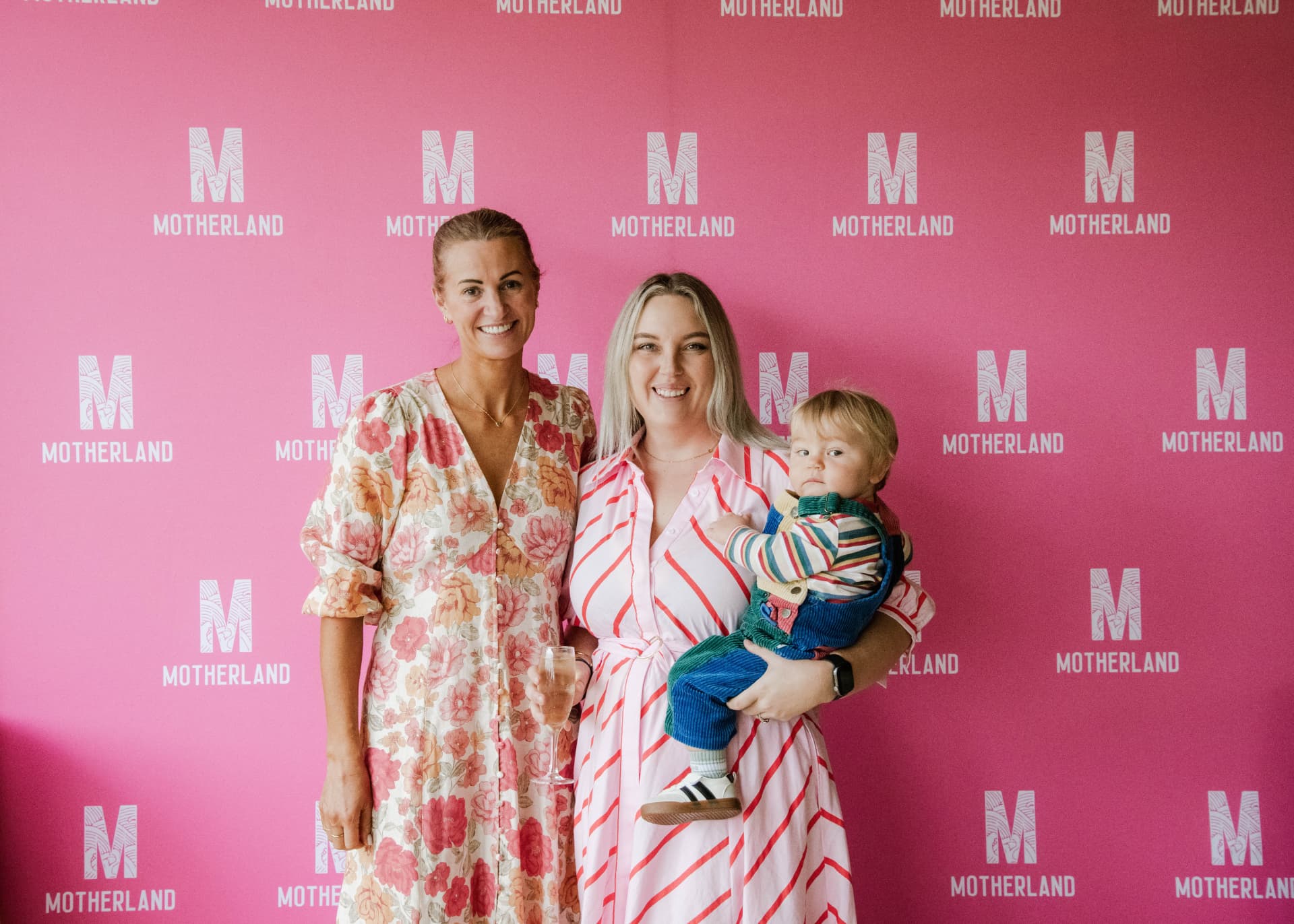 Two mums with a toddler in front of the Motherland backdrop at a community event