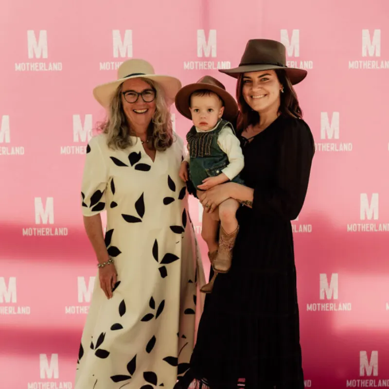 Two women and a toddler smiling in front of a pink Motherland backdrop